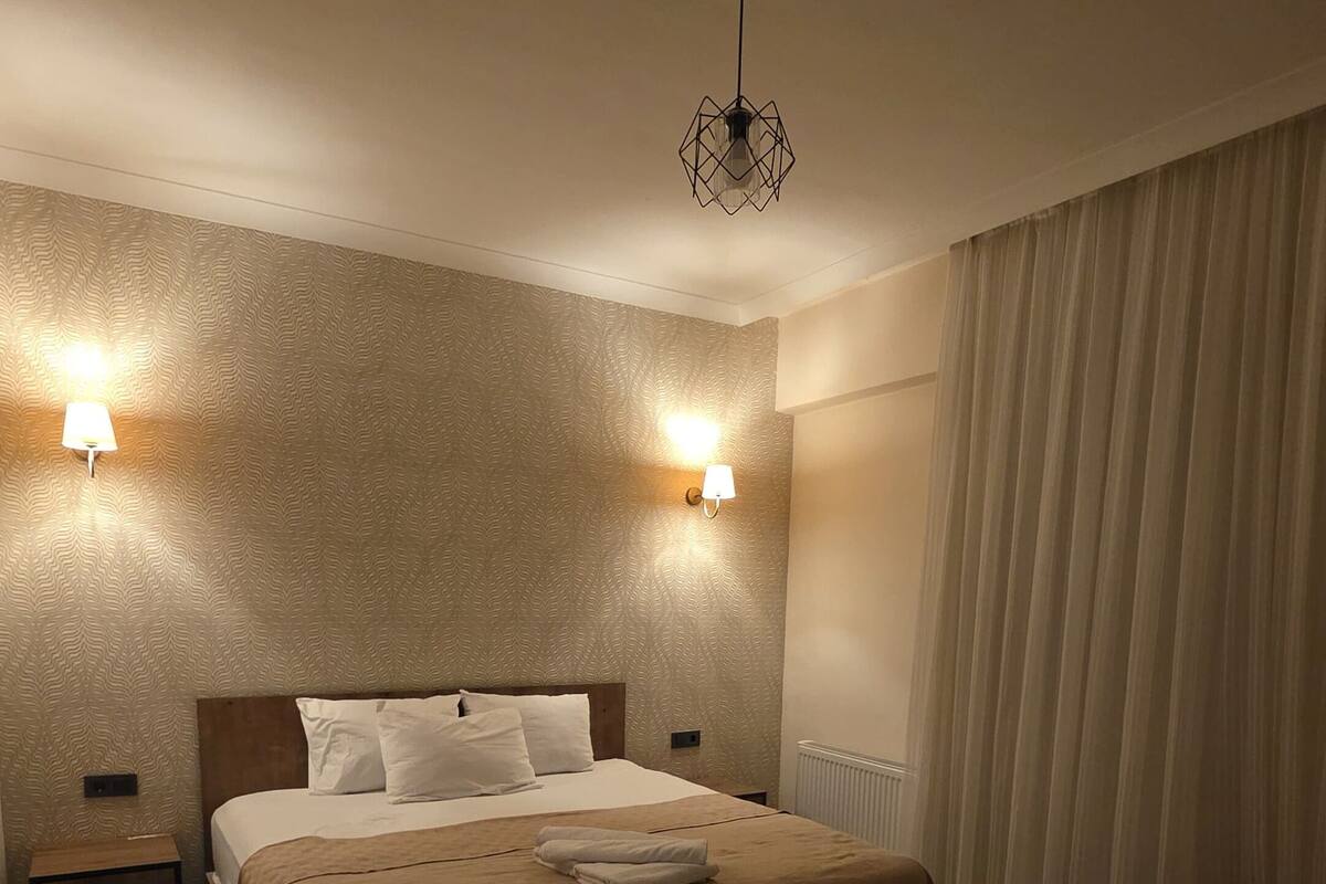 A hotel room with a double bed, two bedside tables, a nightstand, a decorative rug, and a ceiling light fixture.