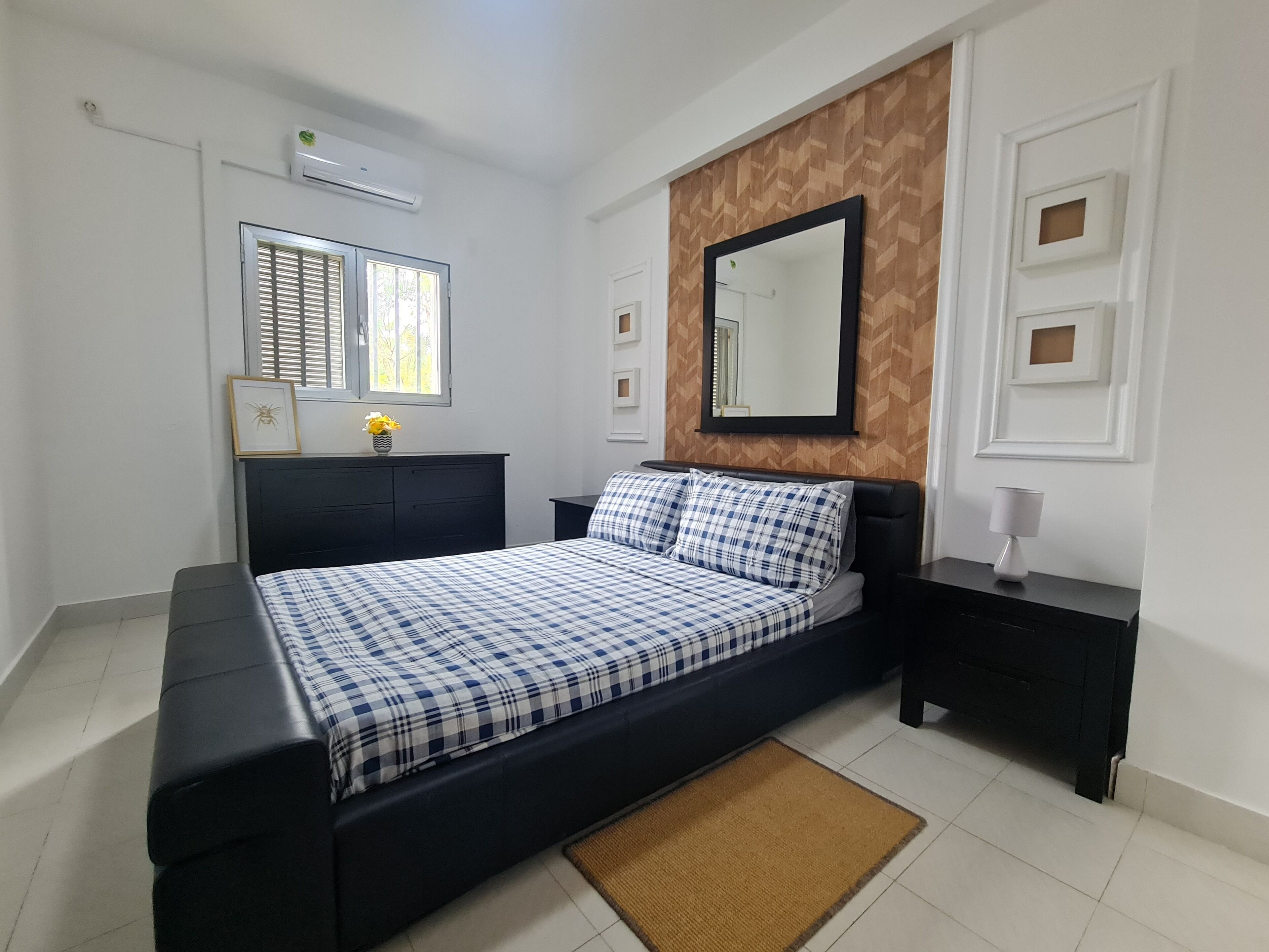 A bedroom with a bed, a bedside table, a nightstand, a mirror, a window with blinds, a wall-mounted air conditioner, and a framed picture on the wall.