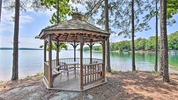 A wooden gazebo with a roof, surrounded by trees, overlooking a lake.