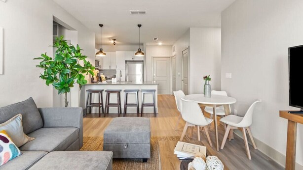 A modern apartment with a living room, dining area, and kitchen. There is a gray sofa, a dining table with white chairs, and a kitchen with a refrigerator and pendant lights.
