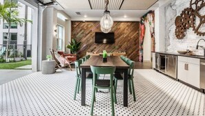 A modern dining area with a wooden table and green chairs, a hanging light fixture, and a kitchen area with white cabinets and a stainless steel sink.
