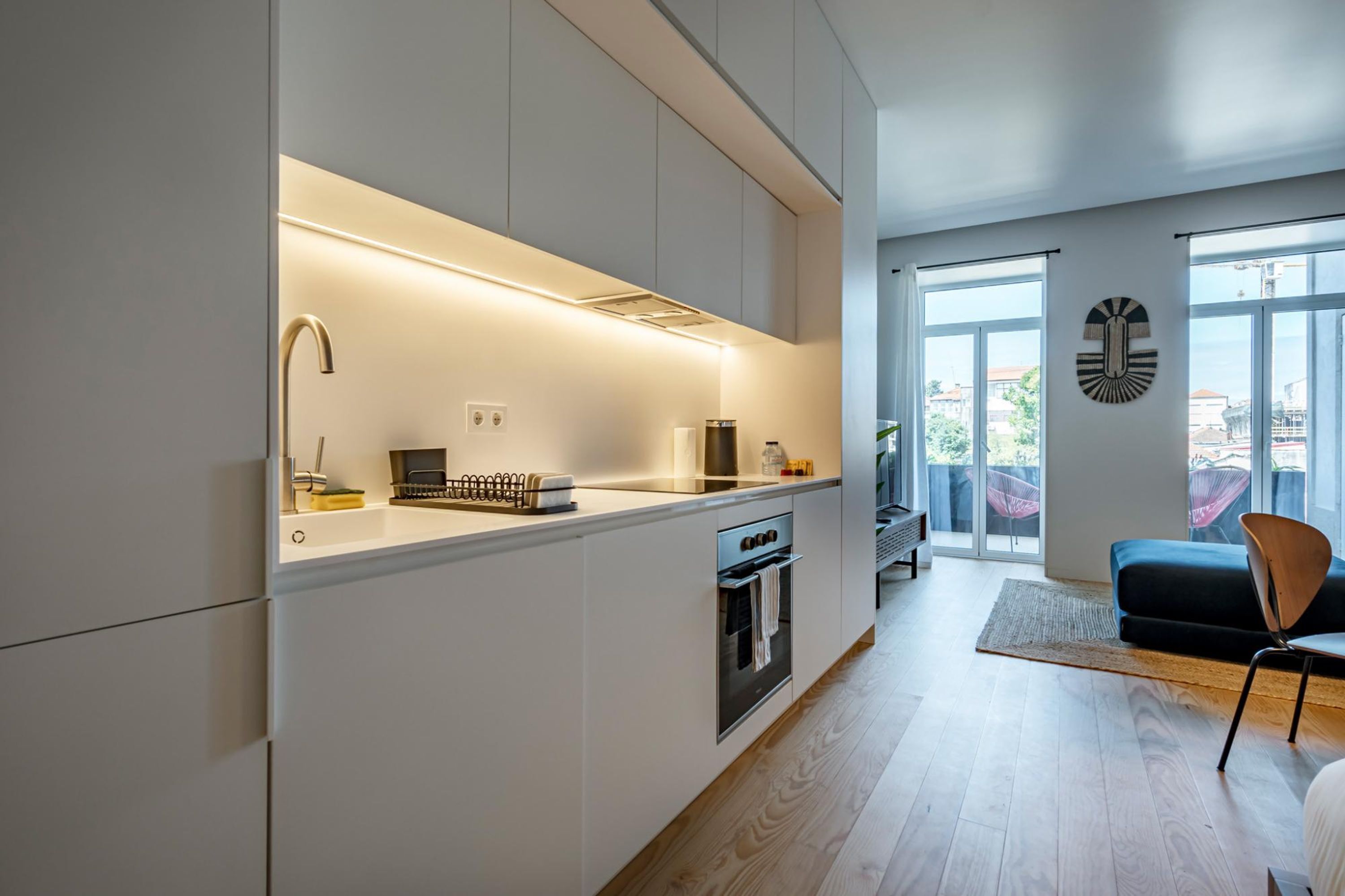 A modern kitchen with built-in appliances, a dining area with a dark sofa and a chair, and large windows offering a view of the city.
