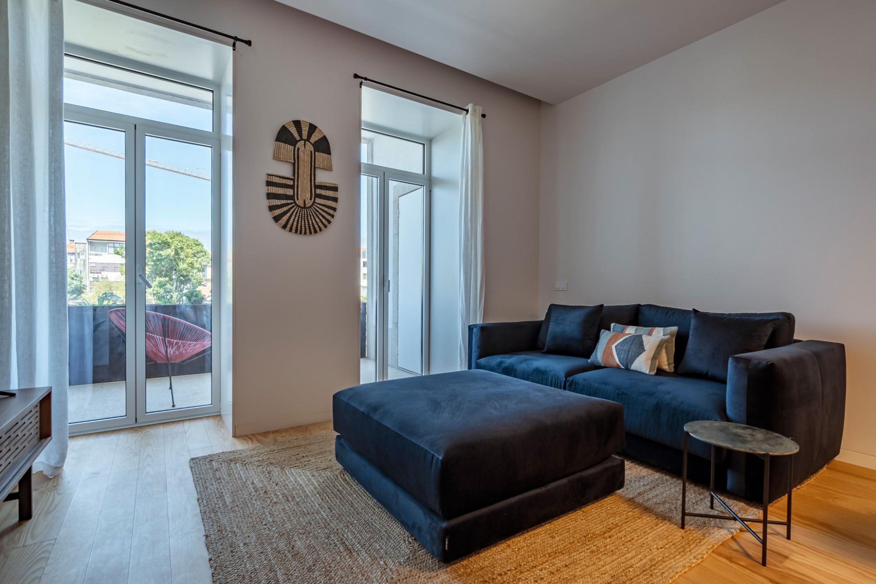 A modern living room with a dark blue sofa, a matching ottoman, and a small side table. There are large windows with a view of the outside, and a decorative wall piece above the sofa.