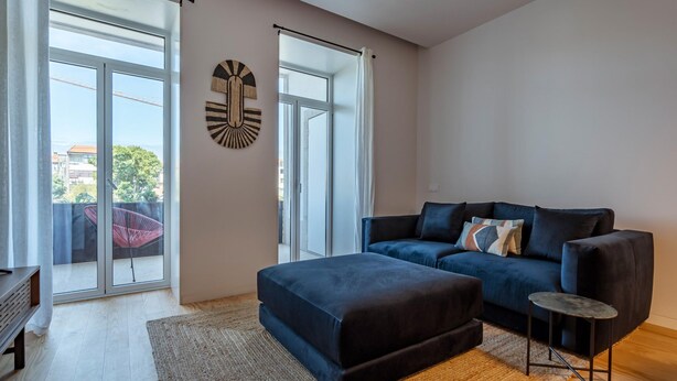 A modern living room with a dark blue sofa, a matching ottoman, and a small side table. There are large windows with a view of the outside, and a decorative wall piece above the sofa. - Nomad s Lux - 1bdr Almada Cool (Porto)