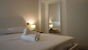 Un lit bien fait, avec du linge de lit blanc et une serviette enroulée, une lampe de chevet et un grand miroir qui reflète la pièce.