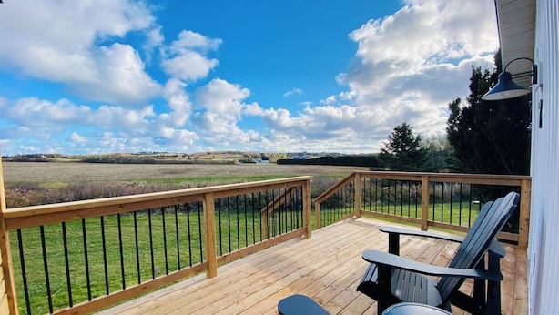 Une terrasse en bois avec une rambarde, des meubles d’extérieur et une vue sur un champ verdoyant et des collines lointaines, le tout sous un ciel partiellement nuageux.