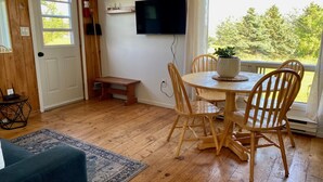 Une pièce au plancher de bois, avec une table et des chaises, un téléviseur fixé au mur et une fenêtre donnant sur des arbres.