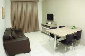 A modern living room with a brown leather sofa, a wooden dining table with four chairs, a television on a stand, and a large window with beige curtains.