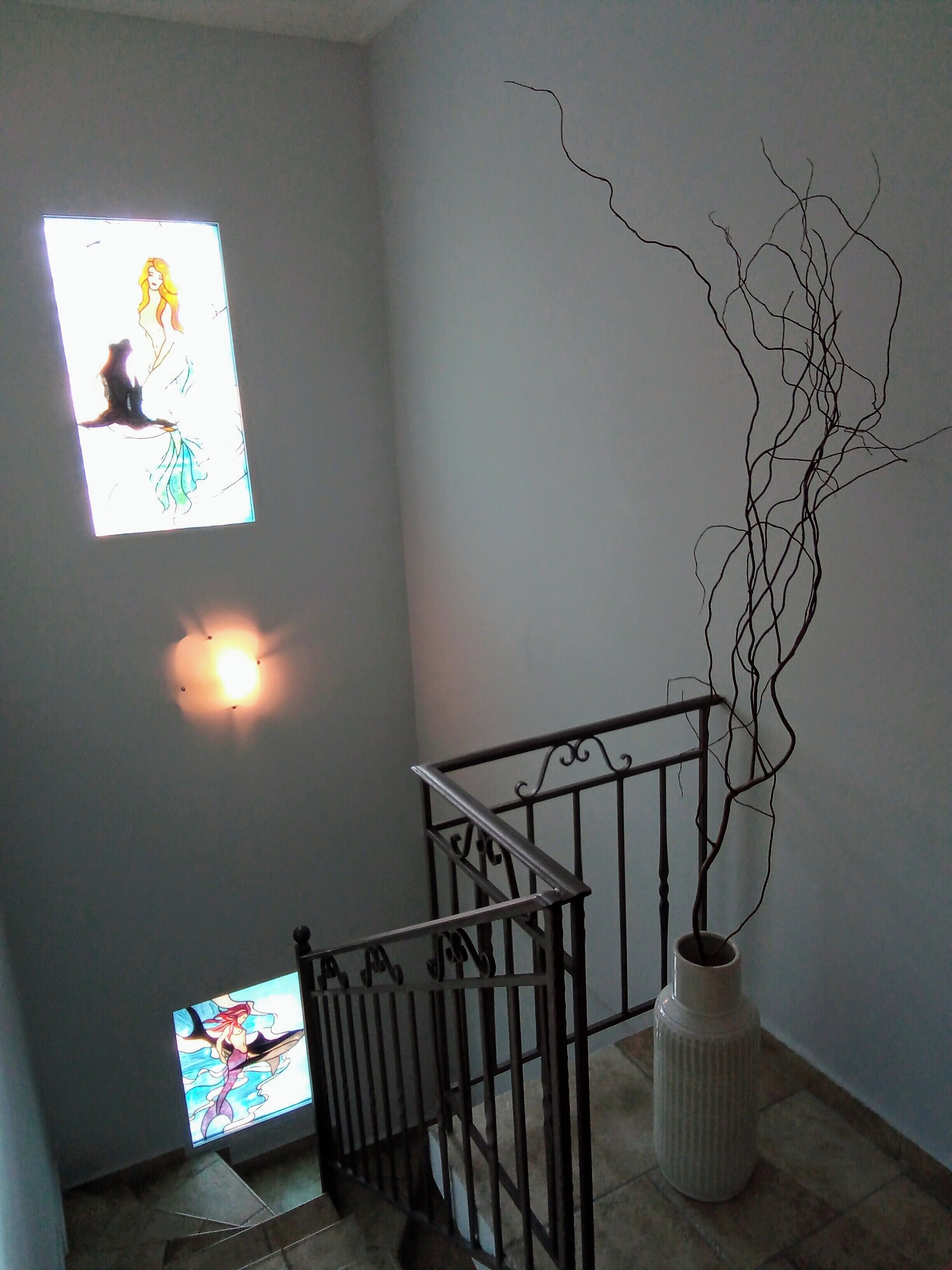 Un escalier avec une rampe en métal, un luminaire fixé au mur et un vase décoratif orné de branches.