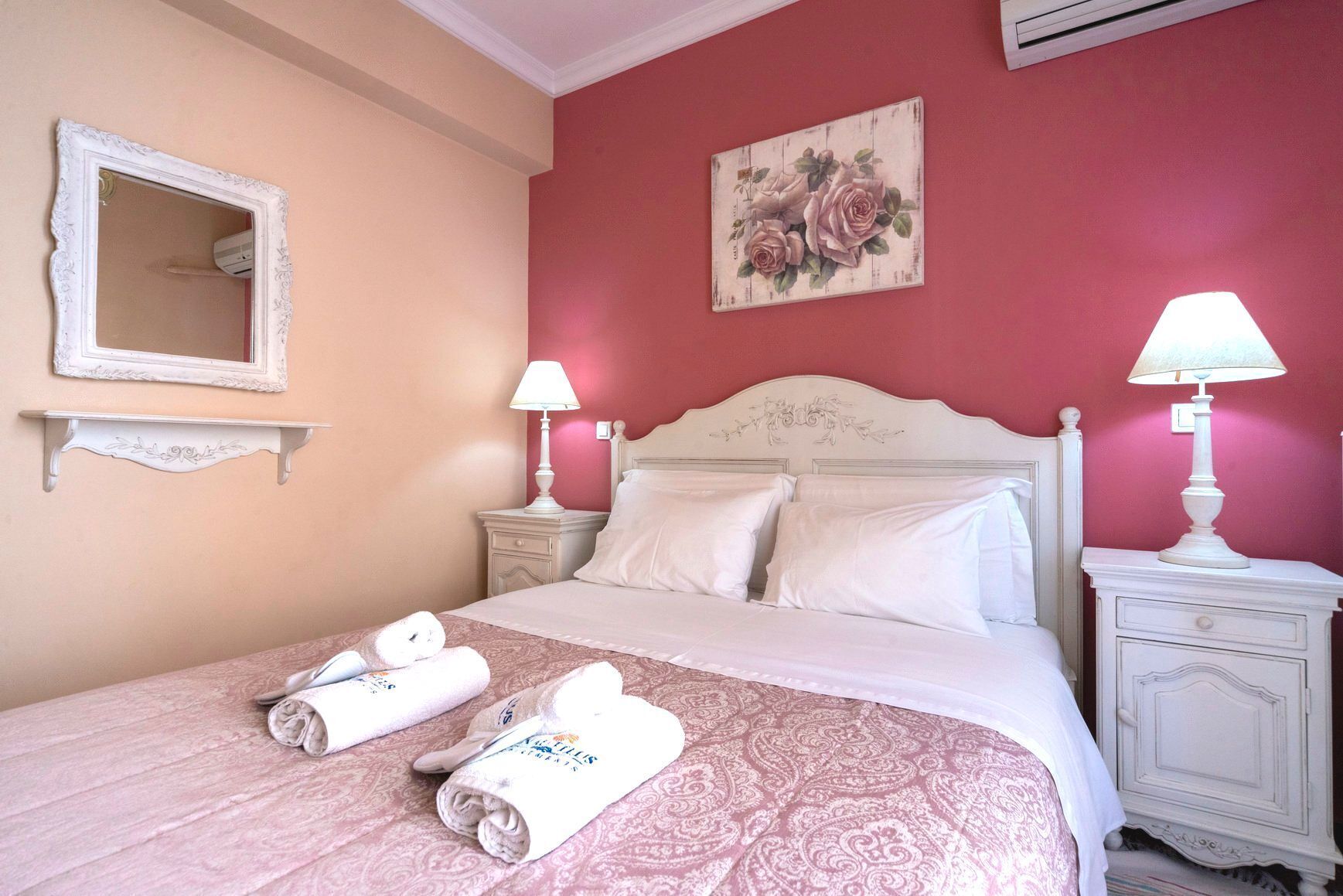 A neatly made bed with pillows and rolled towels, a bedside table with a lamp, a wall-mounted mirror, and a floral artwork above the bed.