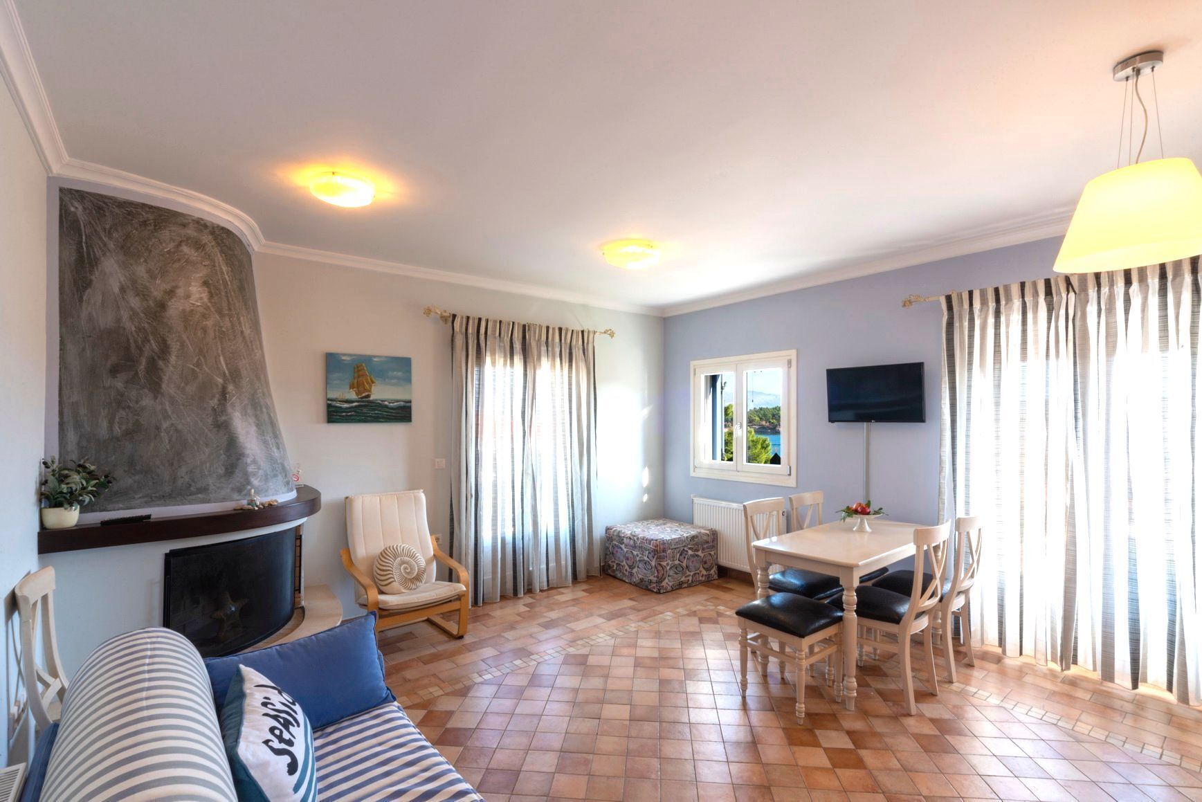 Un salon avec une cheminée, un coin repas avec une table et des chaises, un canapé avec des coussins à rayures et une vue par la fenêtre.