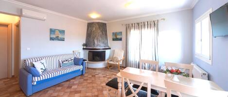 Una sala de estar con sofá, chimenea, mesa de comedor con sillas y televisor montado en la pared.