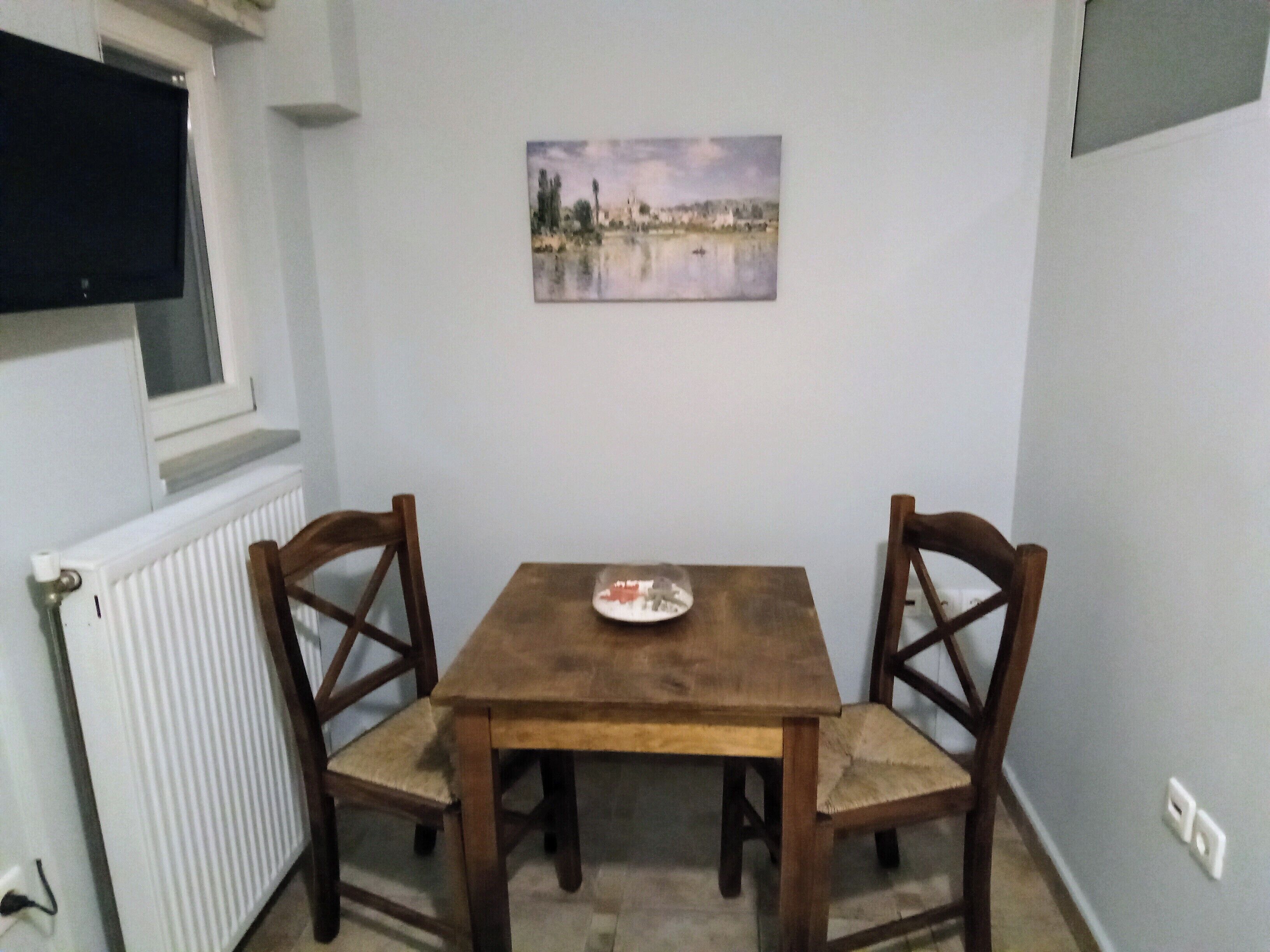 A small dining area with a wooden table and two chairs, a white radiator, and a painting on the wall.