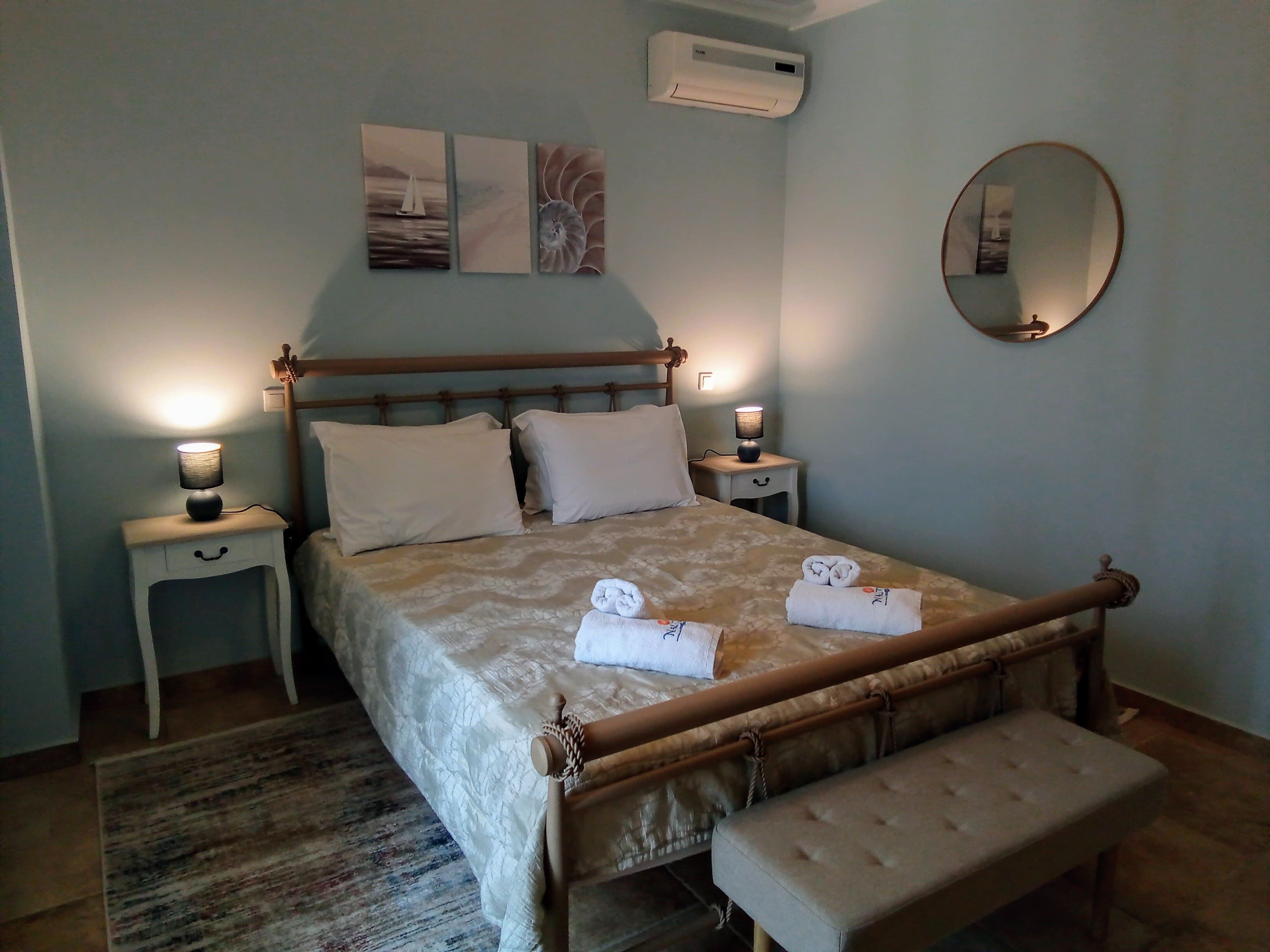 Ein ordentlich eingerichtetes Schlafzimmer mit Bett, Nachttischlampen, einem Nachttisch, einer Bank, einem Spiegel und Kunstwerken an der Wand.