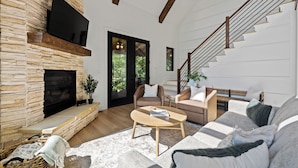 A modern living room with a stone fireplace, a flat-screen TV, a staircase, and a large sofa. - BRAND NEW 4BR 4 BATH & BUNK ROOM MODERN MOUNTAIN LODGE WITH BEAUTIFUL VIEWS (Townsend)