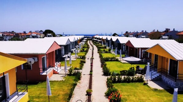 A row of colorful, single-story cabins with a central pathway, surrounded by greenery and a clear view of the sea. - Happy Life Houses (Seferihisar)