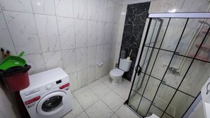 A compact bathroom with a washing machine, toilet, and a glass-enclosed shower area. - Happy Life Houses (Seferihisar)