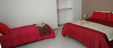 Una habitación con dos camas individuales, cada una con ropa de cama roja y una manta a cuadros. Hay un estante blanco contra la pared y el suelo está cubierto de baldosas.