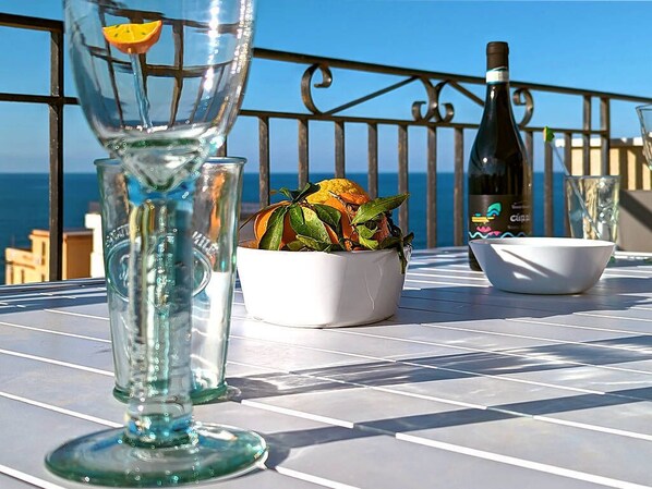 Uma mesa com um copo, uma tigela e uma garrafa, apoiada em um corrimão de varanda com vista para o mar.