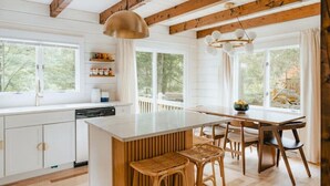 Une cuisine moderne avec des meubles blancs, un îlot central doté de tabourets, des poutres en bois et de grandes fenêtres offrant une vue sur l’extérieur.