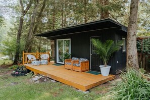 Une petite cabane en bois dotée d’une terrasse, de deux chaises blanches ornées de coussins à motifs et d’un palmier en pot.