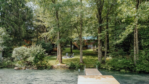 Une jetée en bois s’étendant dans un lac, entourée d’arbres denses et avec une maison visible au loin.