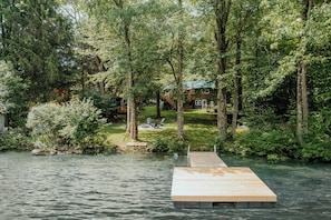 Une jetée en bois s’étendant dans un lac, entourée d’arbres denses et avec une maison visible au loin.