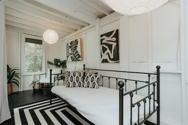 Une chambre à coucher avec un lit en métal noir, du linge de lit blanc, un tapis rayé noir et blanc et des œuvres d’art abstraites accrochées au mur.