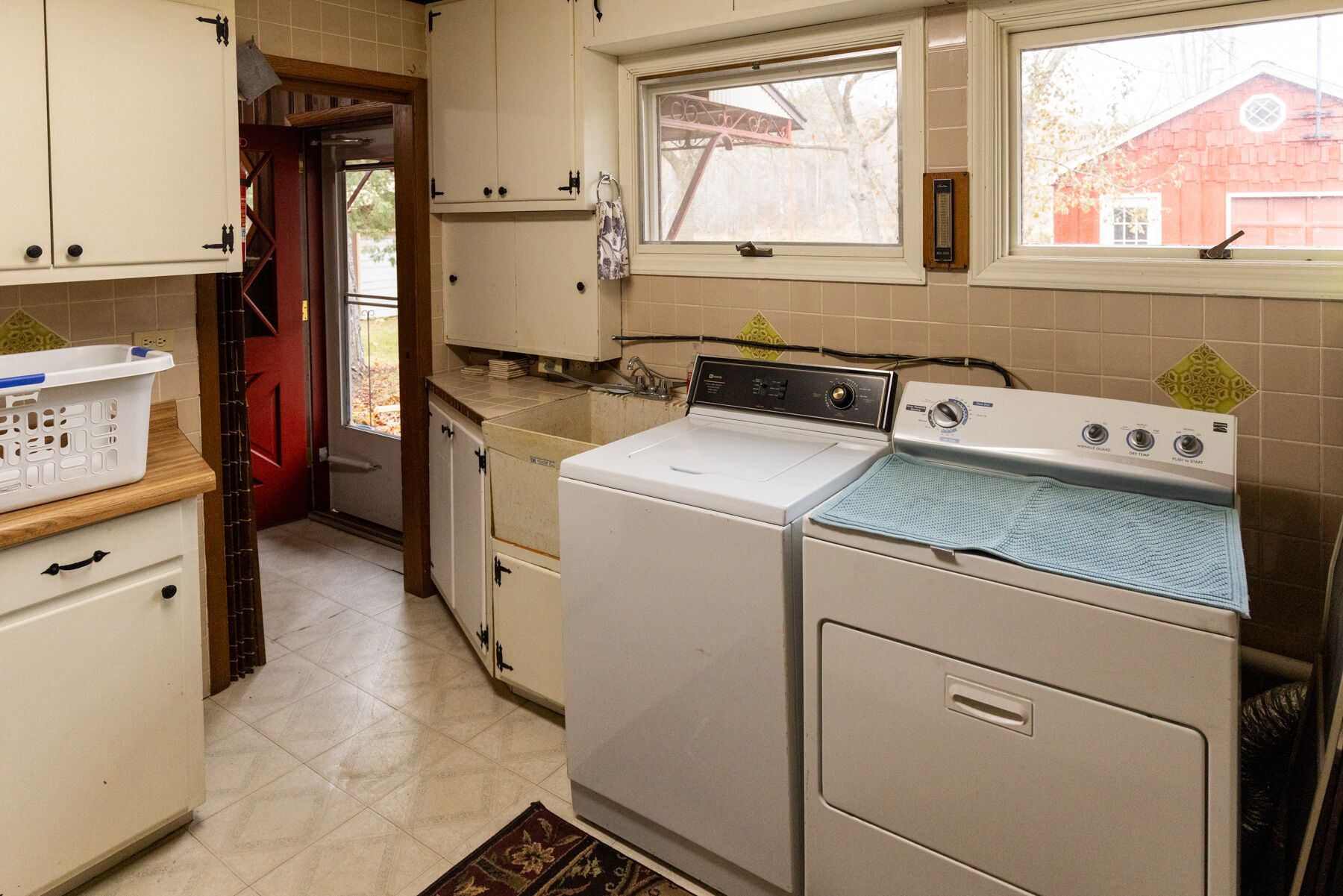 Een wasserette met een voorlader wasmachine en droger, houten kastjes, een raam met uitzicht op een rood gebouw, en een deur naar een andere ruimte.