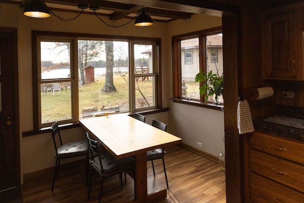 A dining area with a wooden table and chairs, large windows with a view of a yard, and wooden cabinetry.