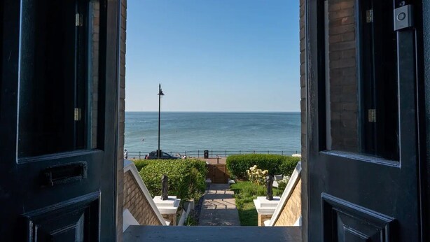 Blick aus einem Fenster auf das Meer bei blauem Himmel, eine Straßenlaterne und ein Weg zu einem Gebäude.