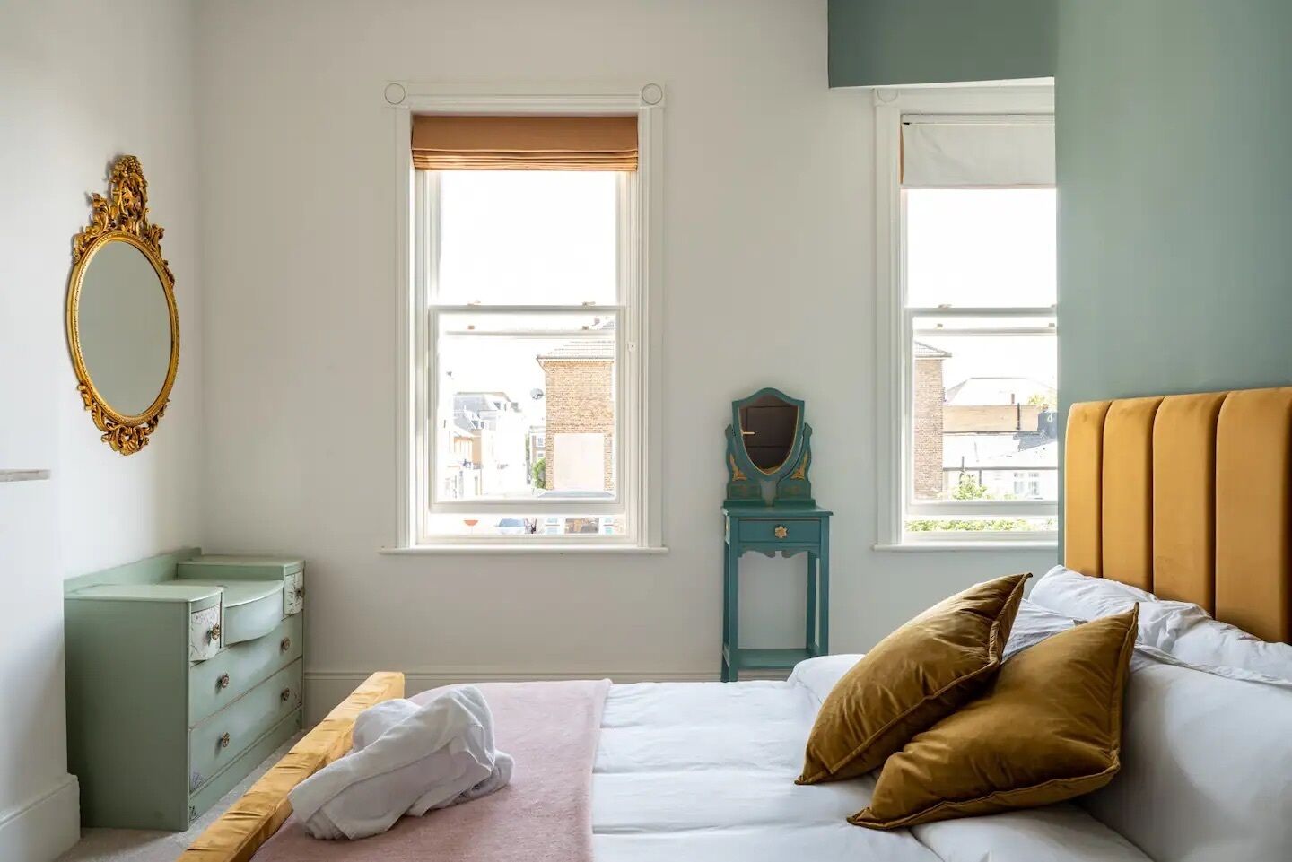 A bedroom with a bed, a dresser, a vanity with a mirror, and two windows with blinds.