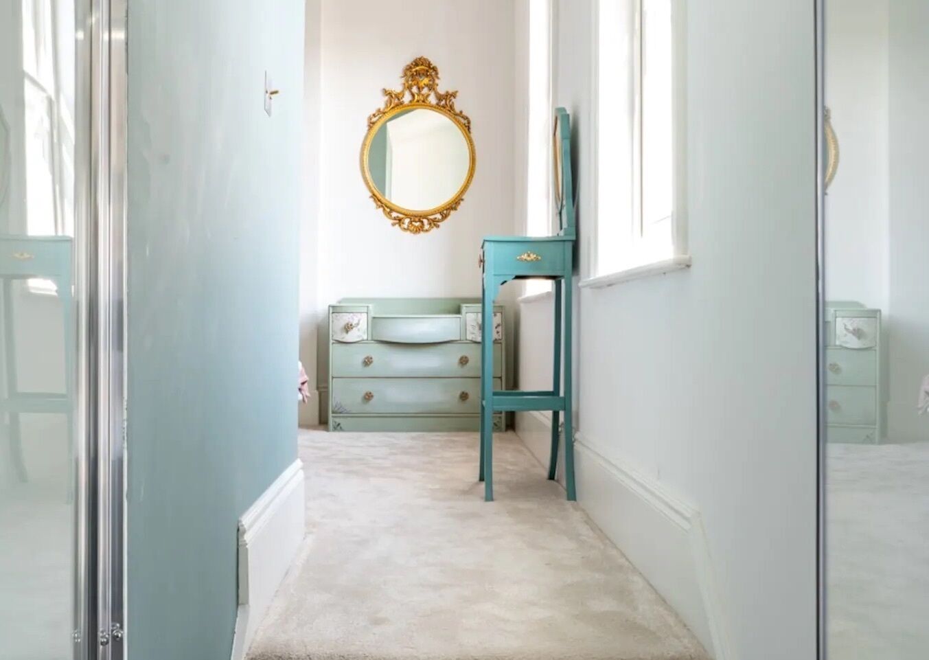 A hallway with a light blue wall, a green dresser with drawers, a mirror with an ornate frame, and a carpeted floor.