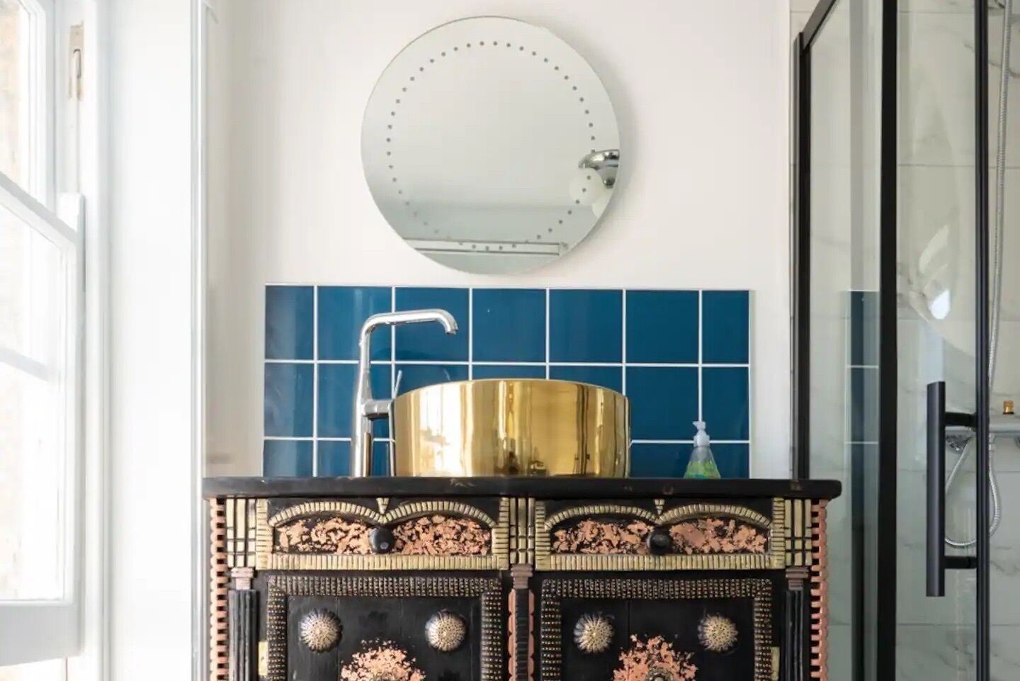 A bathroom with a round mirror, a gold sink, and blue tiles.