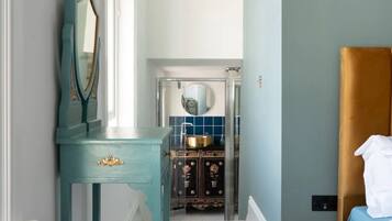 Une pièce avec une coiffeuse de couleur turquoise, un lit avec une tête de lit blanche et une salle de bain visible à travers une porte ouverte.