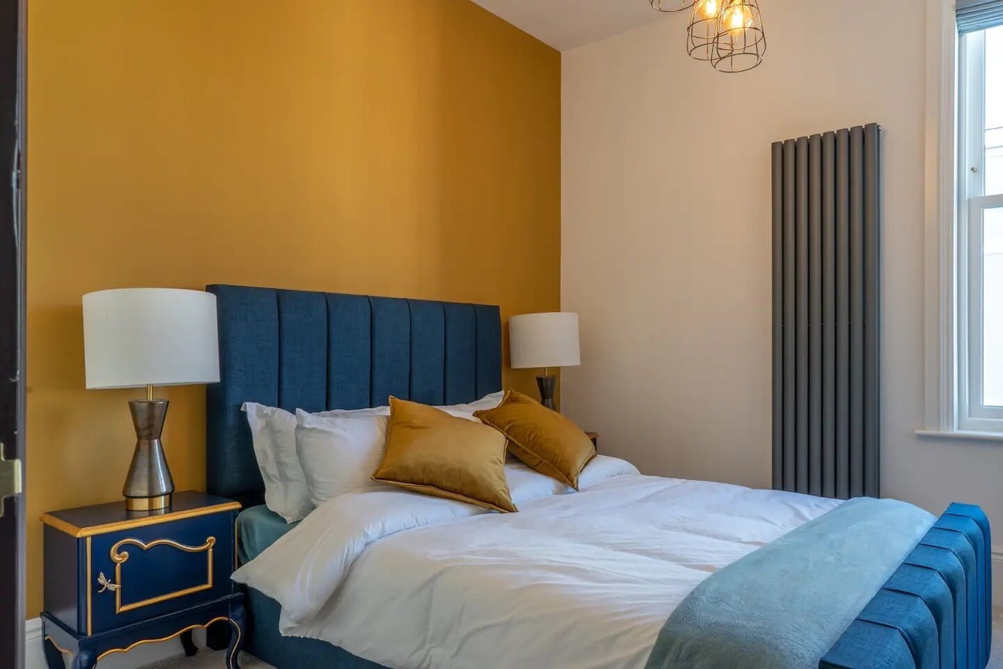 Een slaapkamer met een blauw hoofdeinde, witte beddengoed en twee bedlampjes.