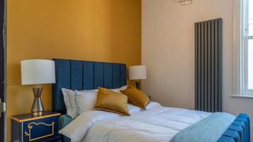Een slaapkamer met een blauw hoofdeinde, witte beddengoed en twee bedlampjes.