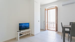 A modern living room with a flat-screen TV on a white stand, a dining area with a table and chairs, and a sliding glass door leading to a balcony. - Chez Isabella by Wonderful Italy (Noicattaro)