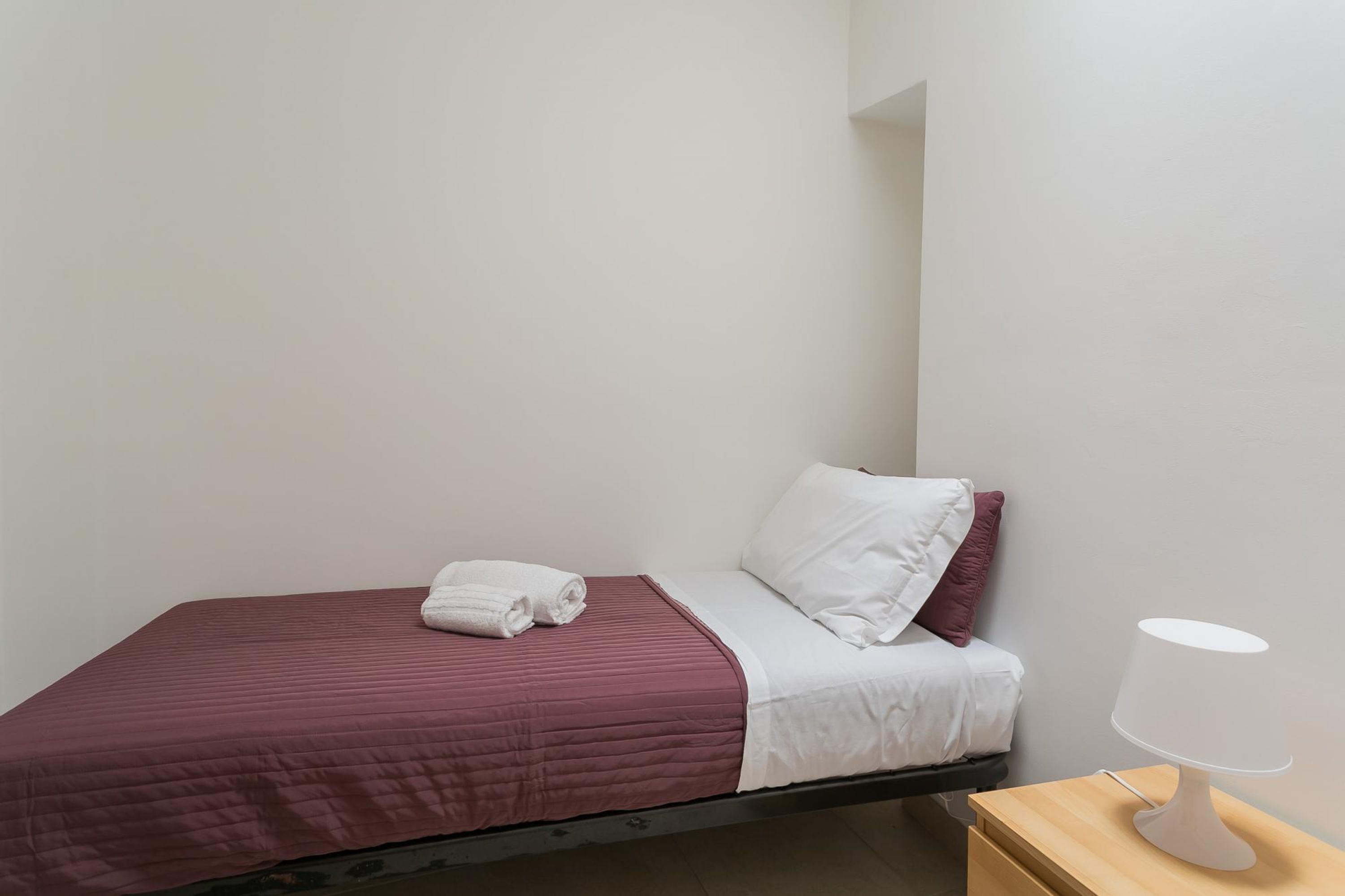 Een eenpersoonsbed met een rode sprei, witte lakens en een wit kussen. Een witte lamp staat op een houten nachtkastje.
