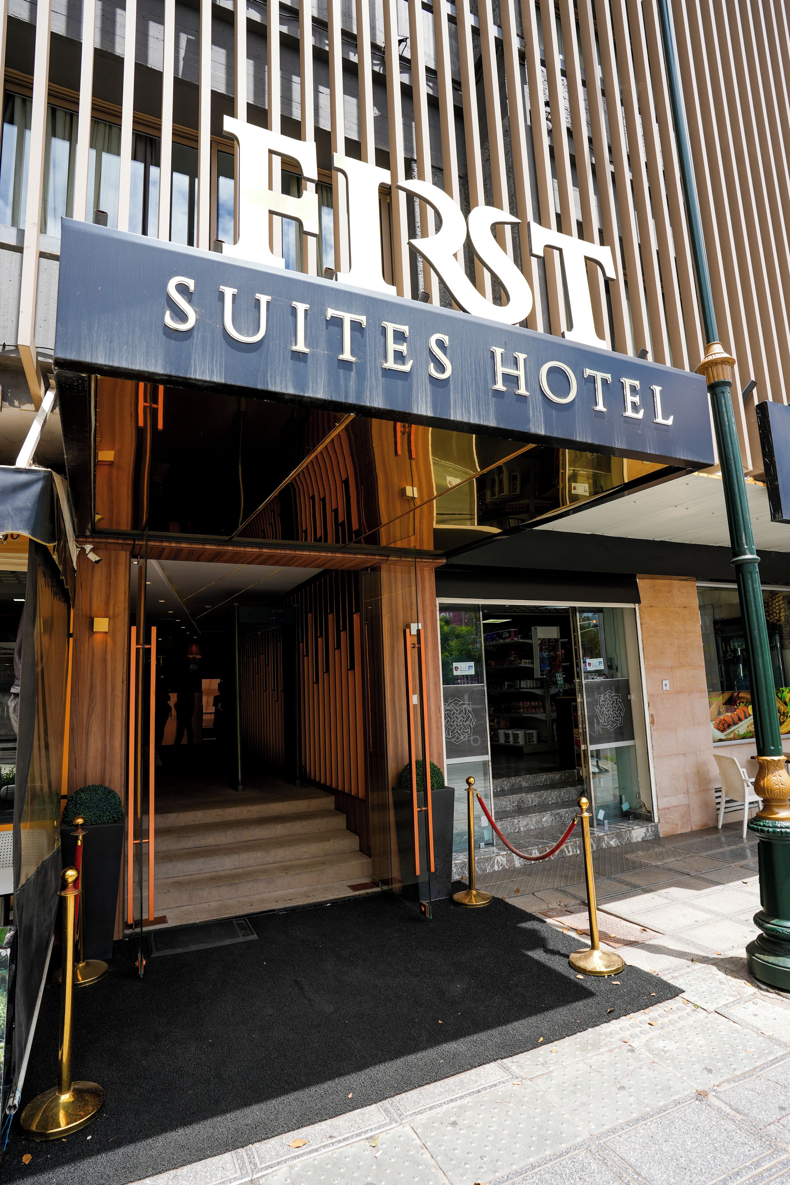 De ingang van Hotel First Suites, met een statig houten gevel, een zwart tapijt en een rode lintbarrière.