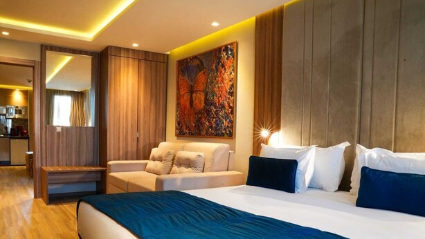 A modern hotel room with a large bed, a sofa, a desk, and a dining area visible through an open door.