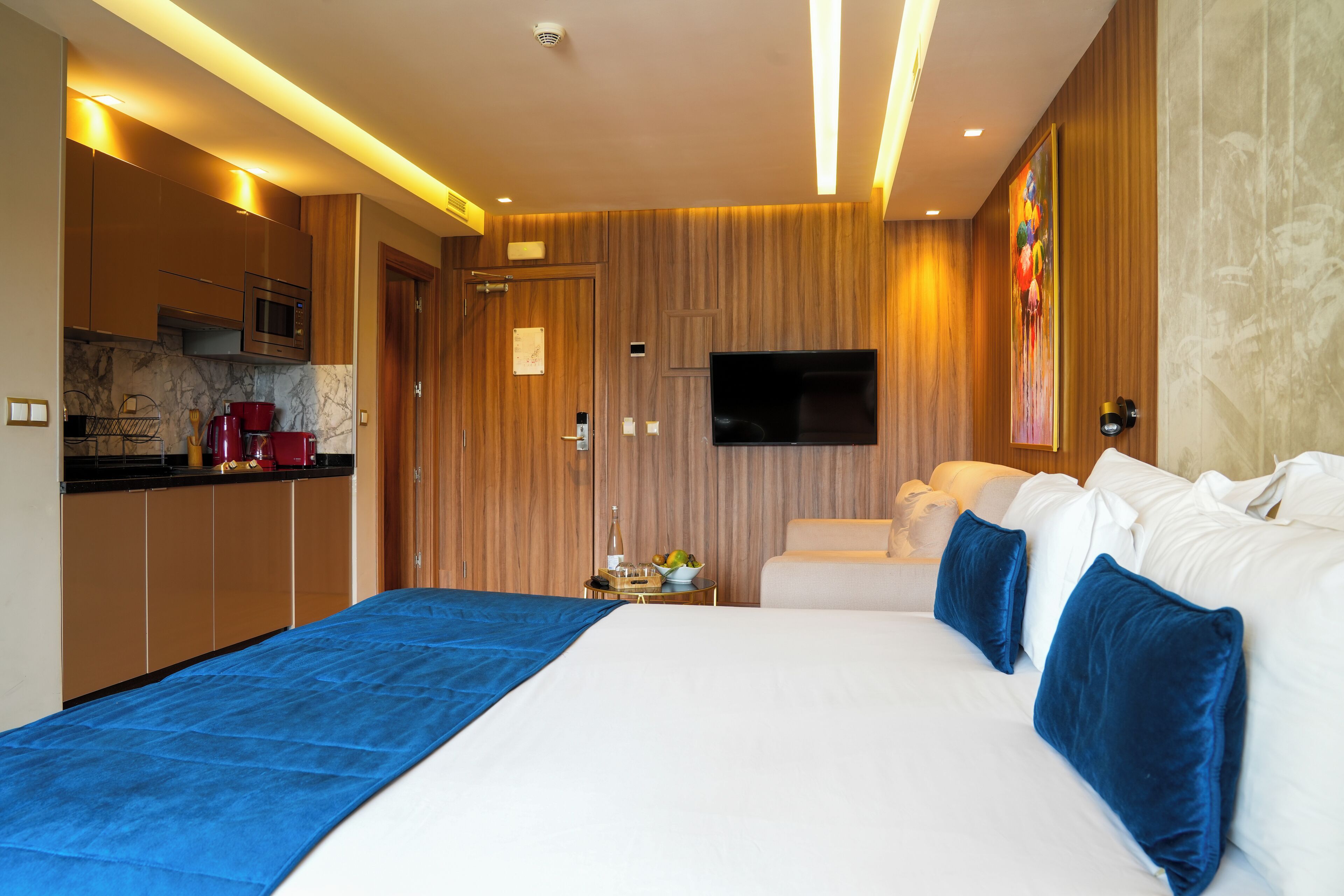 A modern hotel room with a large bed, a TV, a small kitchen area, and a wooden accent wall.