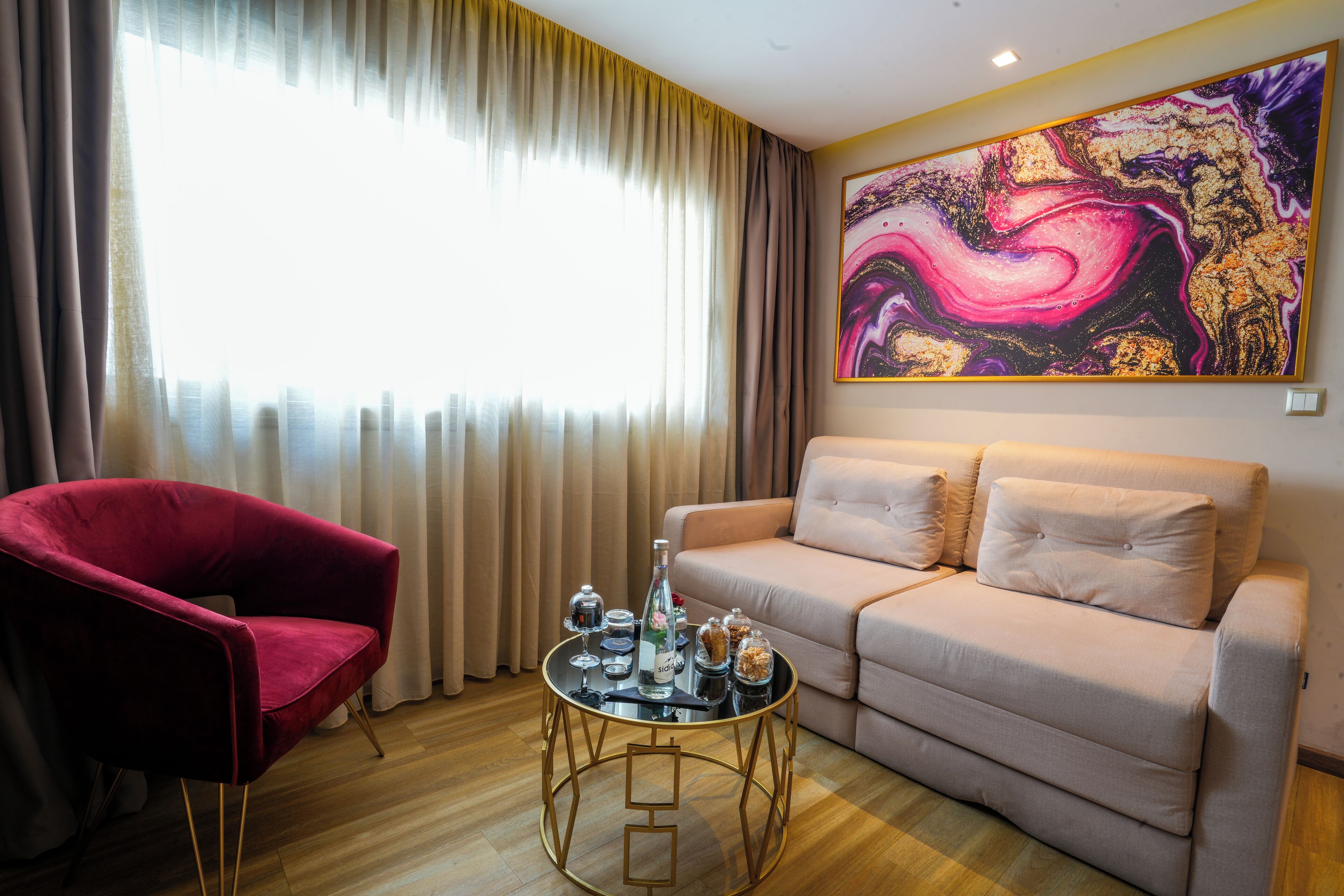 A living room with a red chair, a beige sofa, a round coffee table, and a large abstract painting on the wall.
