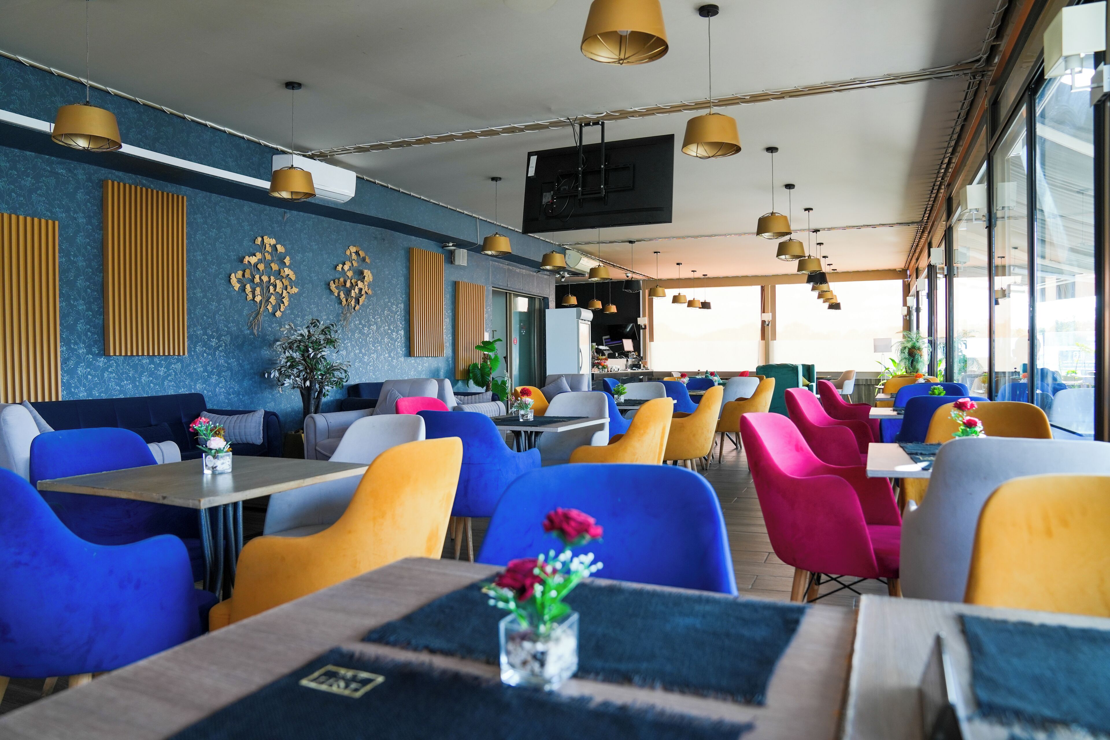 Un restaurante con sillas y mesas coloridas, un televisor en la pared y ventanas amplias.