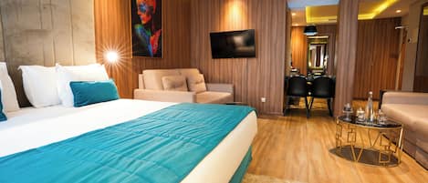 A modern hotel room with a large bed, a sofa, a dining table, and a TV mounted on the wall.