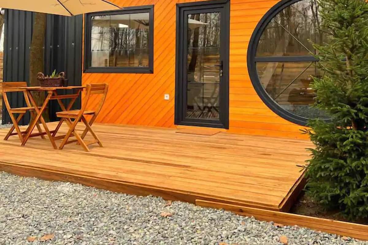 Uma cabana moderna com um deck de madeira, uma área de jantar com mesa e cadeiras, e um janelão circular.