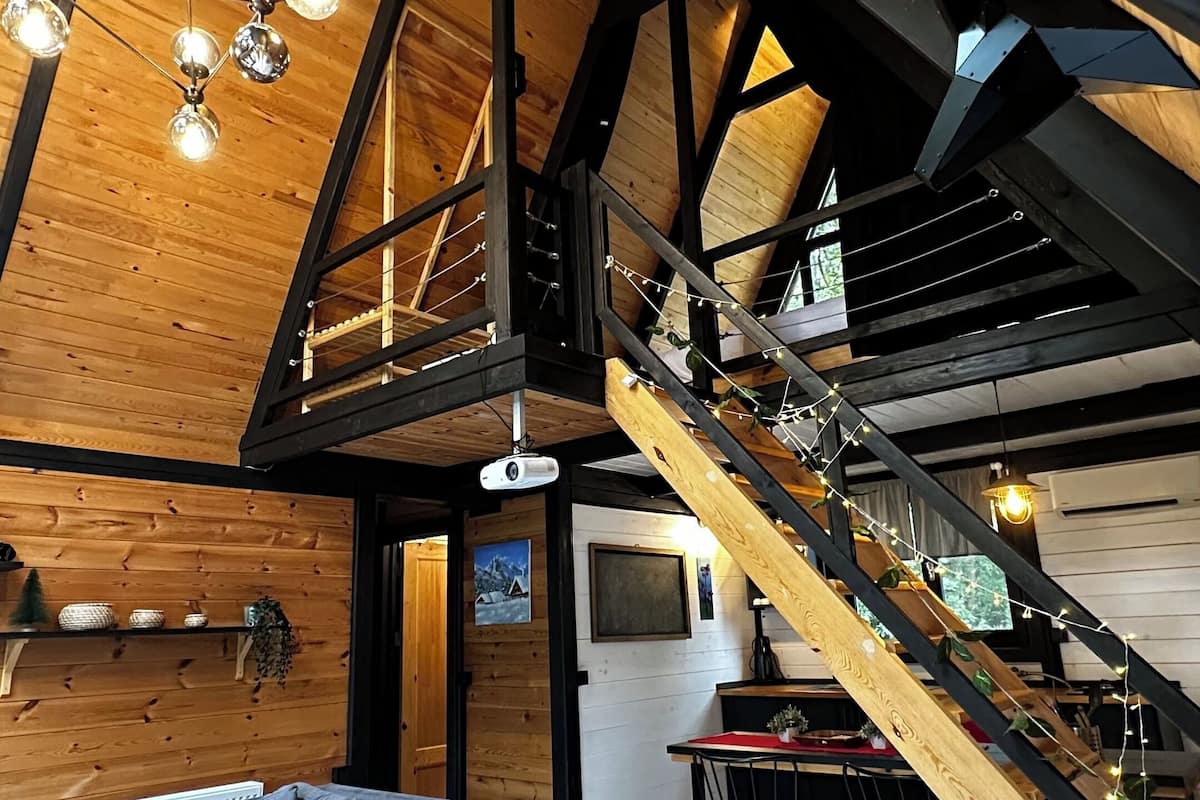 Um amplo loft com teto alto, vigas de madeira aparentes, uma escadaria central, um espaço de estar aconchegante com um sofá e uma mesa de jantar.