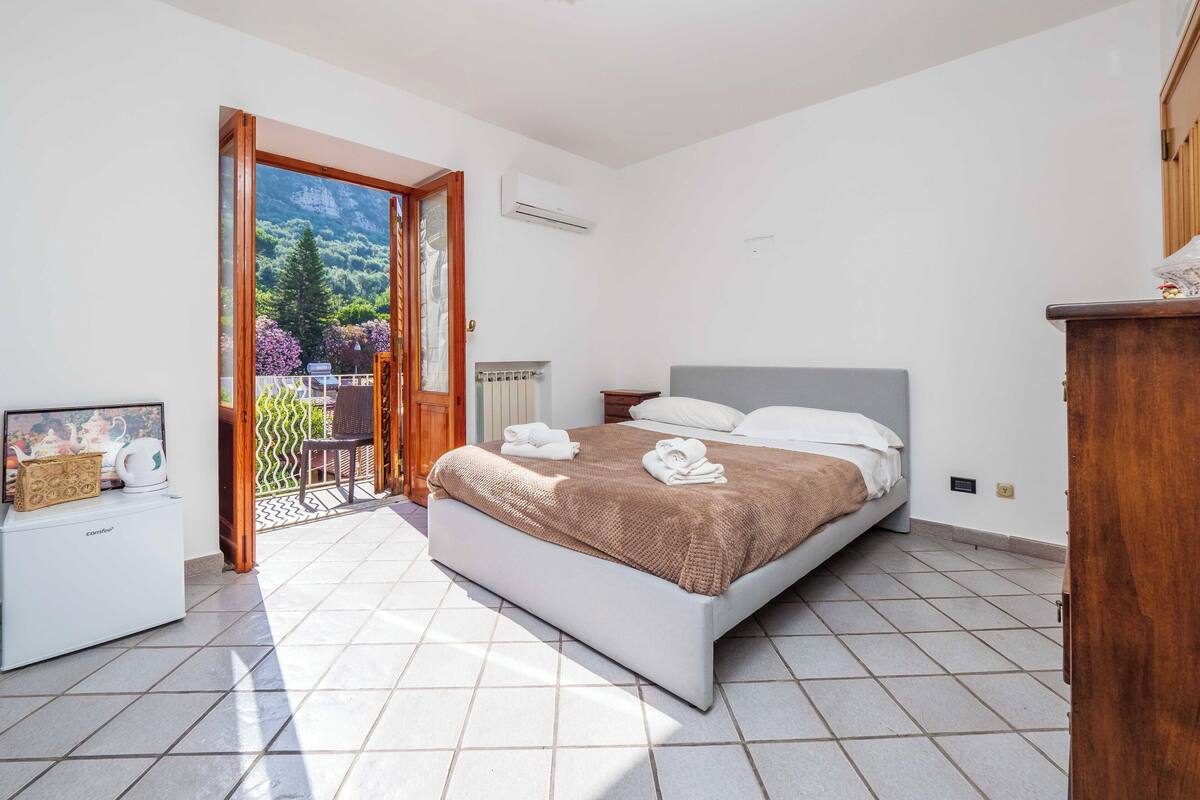 Un dormitorio con cama, mesita de noche, cómoda y vistas a un balcón con plantas.