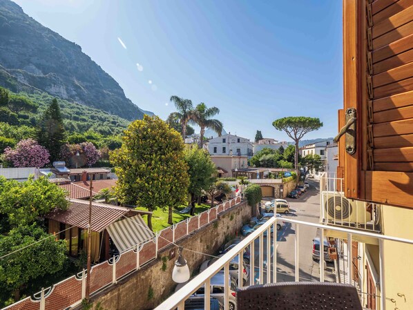 A balcony with a view of a street, a building with a red roof, and a mountain in the background. - Maison Rosa (Meta)