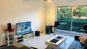 Un salon moderne doté d’un téléviseur à écran plat, d’une table basse, d’un canapé et d’une grande fenêtre donnant sur l’extérieur.